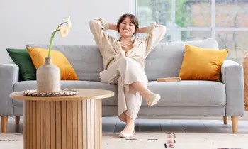 Eine Frau sitzt glücklich, mit den Händen verschränkt hinter ihrem Kopf, auf einem grauen Sofa in einem Wohnzimmer.&nbsp;