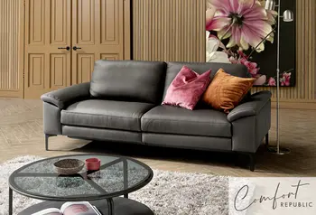 Elegantes Sofa von Comfort Republic in dunkelgrauem Leder, dekoriert mit farbigen Kissen in Rosa und Orange. Im Hintergrund ein stilvoll eingerichtetes Wohnzimmer mit Blumenmotiv und Holzvertäfelung.