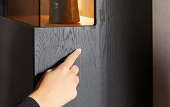 Nahaufnahme einer mattschwarzen Wohnwand mit feiner Holzstruktur – eine Hand demonstriert die grifflose Push-to-Open-Funktion, modernes Design trifft auf innovative Technik für stilvolle Wohnzimmermöbel.