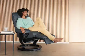 Frau entspannt sich in einem Stressless Mayfair Sessel in der Farbe Paloma Shadow Blue mit Classic-Untergestell in Schwarz. Neben dem Sessel steht ein runder Beistelltisch mit einer Tasse und einem Buch.