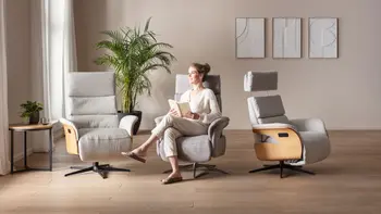 Eine Frau sitzt auf dem mittleren von drei nebeneinander stehenden hellgrauen Relaxssessel mit Holzverkleidung an den Armlehnen außen.