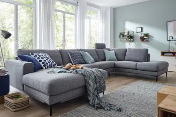 Graue Wohnlandschaft mit einer Kopfstütze in einem hellen Raum mit großen Fenstern.