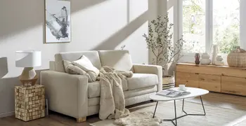 Beiges Zweisitzer Sofa in einem hellen Wohnzimmer, das mit einer Decke und Kissen in beige gemütlich dekoriert ist.