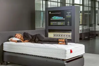 Frau liegt seitlich auf einer unbezogenen Matratze, im Hintergrund ist ein Display zu sehen, auf dem eine Körpervermessung zur Auswahl der perfekten Matratze stattfindet.