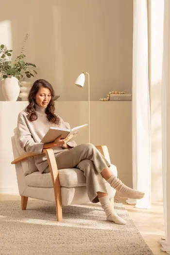 Frau sitzt auf einem beigen Sessel mit Holz Armlehnen und liest ein Buch.