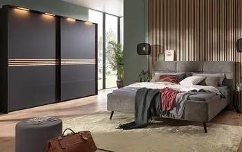 Modernes Schlafzimmer mit grauem Polsterbett und dunklem Schwebetürenschrank mit Holzdekor-Elementen, akzentuiert durch eine Holzlamellenwand, warmes Lichtkonzept und gemütliche Textilien für eine stilvolle und einladende Atmosphäre.