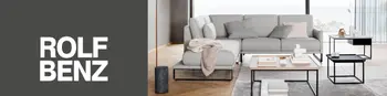 Hellgraues Ecksofa von Rolf Benz mit dünnen schwarzen Standfüßen. Davor und daneben Beistelltische. Links auf einer grauen Fläche das Rolf Benz Logo.&nbsp;