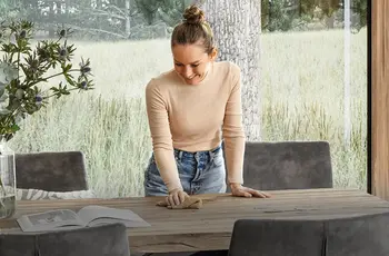 Lächelnde Frau steht über einem Esstisch aus Massivholz und wischt mit einem Lappen darüber.