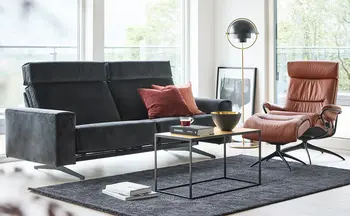 Elegantes Wohnzimmerensemble mit einem schwarzen Sofa, einem braunen Relaxsessel und einem modernen Couchtisch, ergänzt durch stilvolle Dekorationen und eine einladende Beleuchtung.