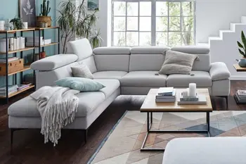 Großes graues Ecksofa mit einer aufgestellten Kopfstütze, dekoriert mit grauen Kissen und grauer Decke in einem hellen Wohnzimmer.