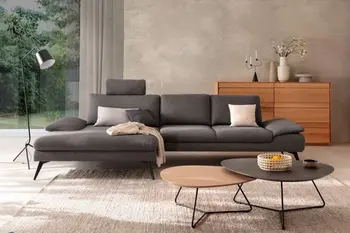 Graues Sofa mit breitem Longchair und schwarzen Beinen in einem gemütlichen Raum mit großem Fenster.