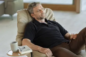 Ein Mann entspannt sich in einem Stressless Reno Sessel in der Farbe Paloma Sand mit Classic-Untergestell aus Eiche. Auf dem eingebauten Beistelltisch liegen ein Buch und eine Tasse.