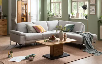 Modernes Wohnzimmer mit grauer Eckcouch, bunten Kissen und quadratischem Couchtisch aus Holz – stilvolle Einrichtung in freundlicher, grüner Raumgestaltung mit großen Fenstern und Tageslicht.