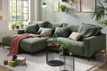 Olivgrünes Ecksofa mit tiefer Sitzfläche und schwarzen Metallfüßen, auf dem Sofa liegen mehrere grüne zum Sofa passende Kissen, sowie zwei beige Kissen und eine rote Decke.