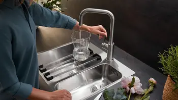 Frau befüllt ein Glas mit Wasser an einer modernen Edelstahlarmatur über einer doppelten Spüle mit Abtropfgitter und floralem Dekor im Vordergrund. Himmlisch Wohnen