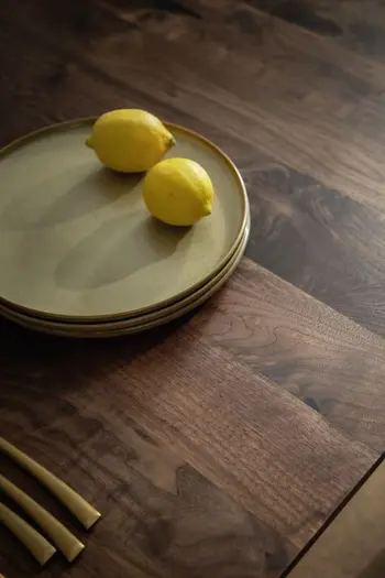 Dunkelbraune Tischplatte eines Esszimmertisches, auf dem ein Tellerstapel steht mit zwei Zitronen oben drauf.