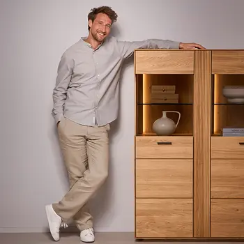 Lächelnder Mann lehnt entspannt an einer modernen Eichen-Vitrine mit stilvoller LED-Beleuchtung – hochwertiges Möbelstück mit Glasfach und Schubladen im skandinavischen Design
