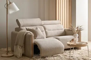 Stressless Emily Sofa mit beige Stoffbezug und Eichenholz-Elementen, ausgestattet mit Relaxfunktion. Dekoriert mit Kissen und Decke, daneben ein runder Beistelltisch mit Vase und Dekorfigur in einem modernen Wohnraum.