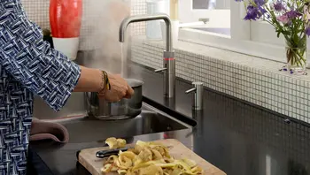 Person füllt mit kochendem Wasser aus einem modernen Wasserhahn einen Kochtopf in einer Küche mit schwarzer Arbeitsfläche und Mosaikfliesen