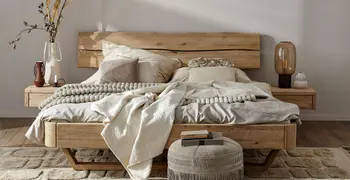 Helles Holzbett mit Nachtisch aus Eichenholz gemütlich dekoriert mit hellen Kissen und Decken und Vasen in Braun und Weiß mit Trockenblumen auf den daneben stehenden passenden Nachttischen.