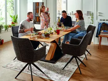 Menschen, die gemütlich an einem Esstisch sitzen und lachen. Eine Frau hat ein Baby auf dem Arm und steht. Der Tisch wird durch besondere, x-förmige Beine gehalten.&nbsp;