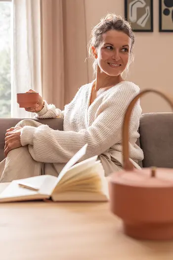 Frau trinkt entspannt eine Tasse Tee und sitzt an Esstisch auf einer gemütlichen Bank.