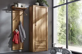 Garderobe in hellem Holz bestehend aus einem Schrank mit Tür und Klapptüre und einer Hutablage mit Kleiderhaken an dem zwei Jacken hängen. 