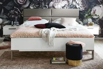 Weißes Bett mit grauem gepolsterten Kopfteil und silbernen Füßen und schwebenden Nachtkonsolen vor einer Wand mit auffälliger Blumentapete.