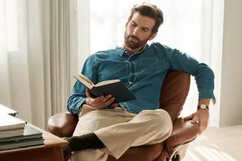 Ein Mann liest ein Buch in einem Stressless Rome Home-Office-Sessel in der Farbe Paloma Dark Henna mit poliertem Untergestell. Im Hintergrund helle Vorhänge und ein moderner Schreibtisch mit Büchern.