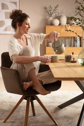 Frau sitzt im Schneidersitz auf einem dunkelgrauen Schalenstuhl mit Holzbeinen und schenkt etwas ein eine Tasse auf dem Tisch ein.