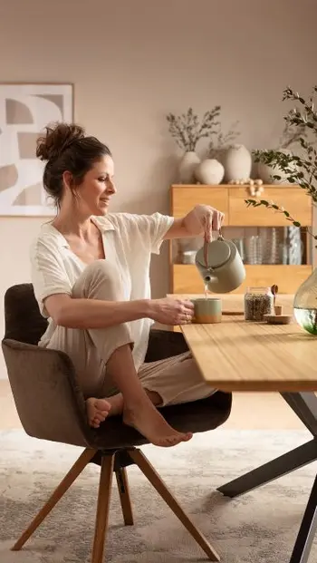 Frau sitzt gemütlich an einem Esstisch aus Massivholz und gießt etwas in eine Tasse.