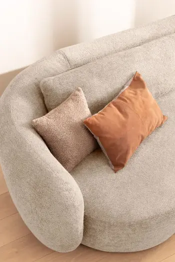 Sitzfläche und abgerundete Armlehne eines cremefarbenen Sofas, auf dessen Sitzfläche zwei Kissen in beige und orange liegen.