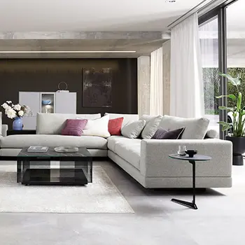 Graue, riesige Wohnlandschaft in einem großen, lichtdurchfluteten Raum. Auf dem Sofa befinden sich viele Kissen in unterschiedlichen Farben, während neben dem Sofa ein kleiner runder Beistelltisch und ein großer eckiger Glastisch stehen.&nbsp;