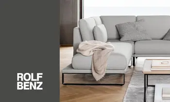 Ein hellgraues Sofa der Marke Rolf Benz, auf der linken Seite befindet sich das Rolf-Benz-Logo&nbsp;auf grauem Hintergrund.