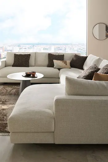 Design Ecksofa Contur Cuneo in beige vor großem Fenster mit Aussicht auf Skyline.