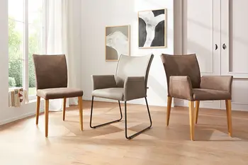 Ein brauner Esszimmerstuhl neben einem grauen Lederstuhl mit Metallgestell und einem braunen Leder Armlehnstuhl mit Holzgestell.