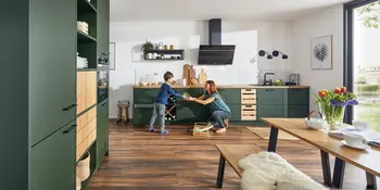 Mutter mit Kind vor gruener Kuechenzeile mit Holzakzenten in moderner offener Kueche.