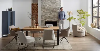 Moderner Massivholz Esstisch, um den herum graue Schalenstühle stehen in einem hellen Raum in dem ein Mann an einer Wand lehnt.