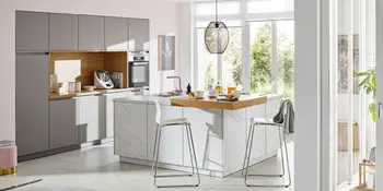 Helle Inselkueche mit quadratischer Kuecheninsel vor grauer Kuechenzeile mit Einbaubackofen.