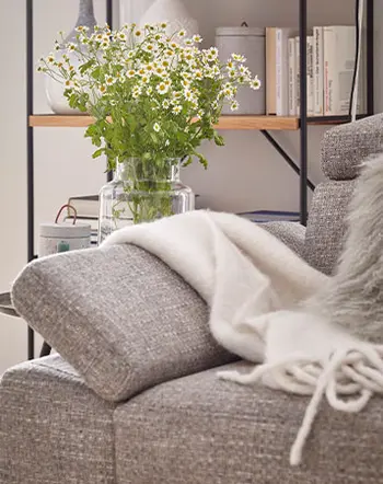 Aufgestellte Armlehne eines grauen Sofas aus Stoff, auf dem eine weiße Decke liegt und im Hintergrund eine Vase mit Blumen zu sehen sind.