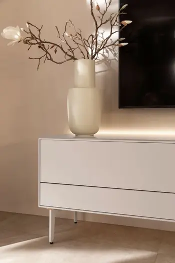 Weißes Lowboard mit Schubladen und weißen Füßen und indirekter Beleuchten an der Rückwand.